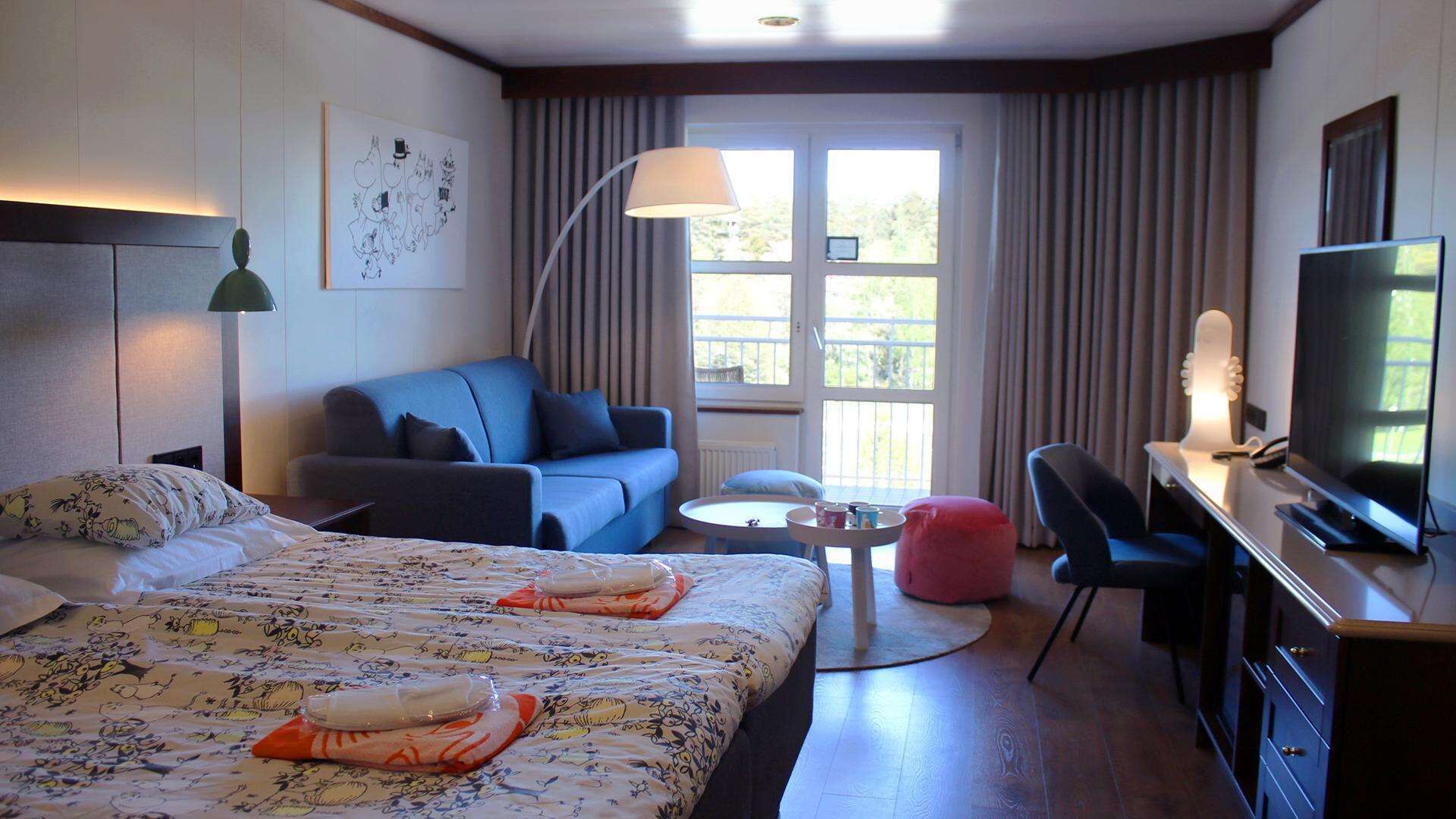 Habitación de hotel con cama, sofá, escritorio con televisor, lámpara y ventana con cortinas.