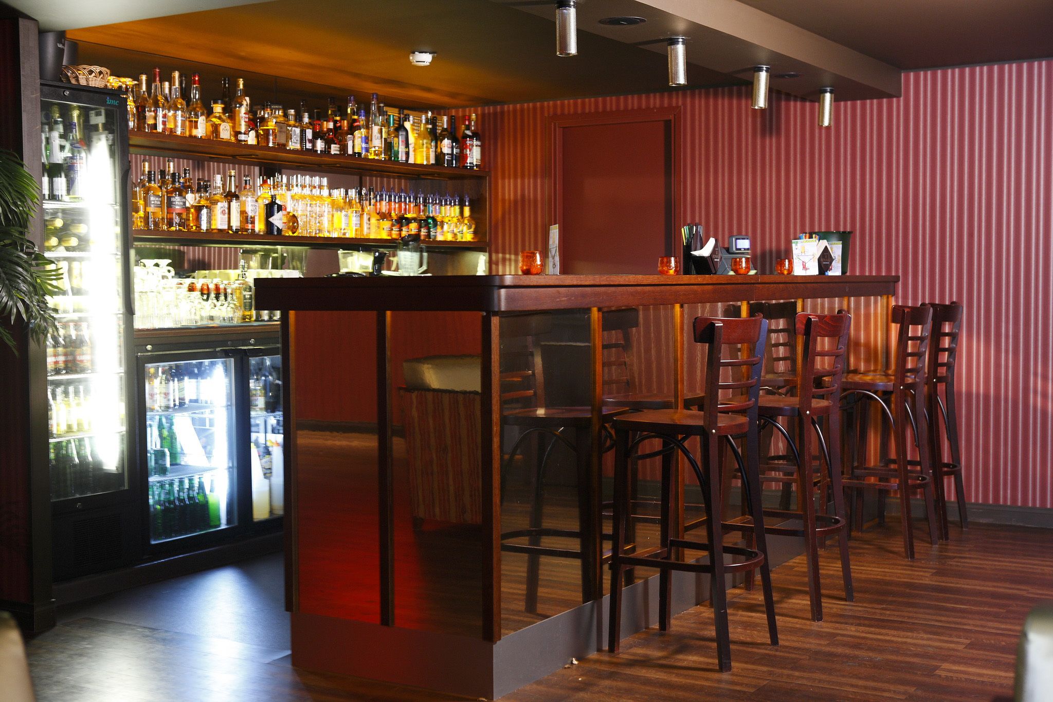 A bar area with a wooden bar counter, multiple stools, and a refrigerator stocked with bottles.