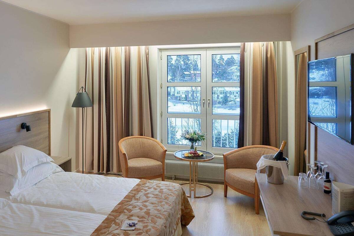 A hotel room with two beds, a desk with a chair, a small table with a vase, and a TV on the wall.
