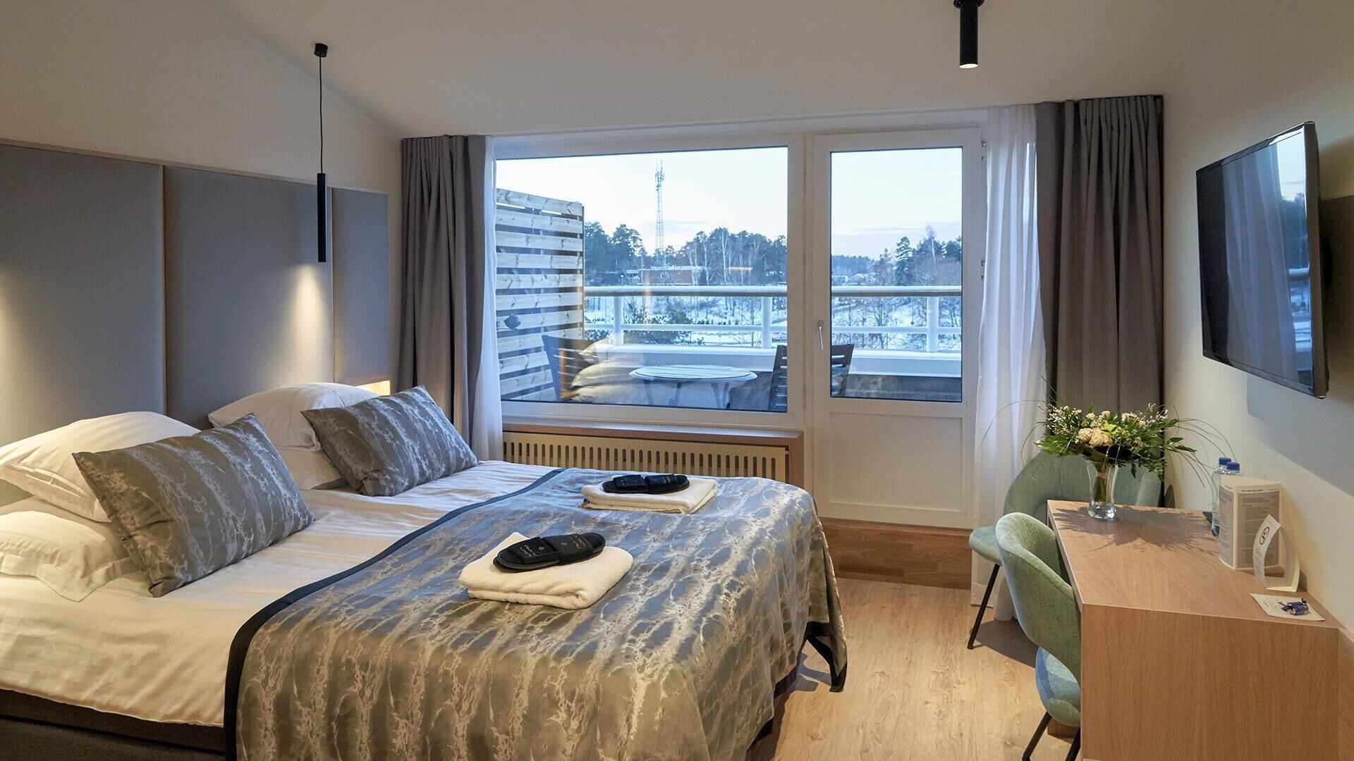 A hotel room with a bed, a desk, a chair, a TV, and a window with a view of buildings and trees.