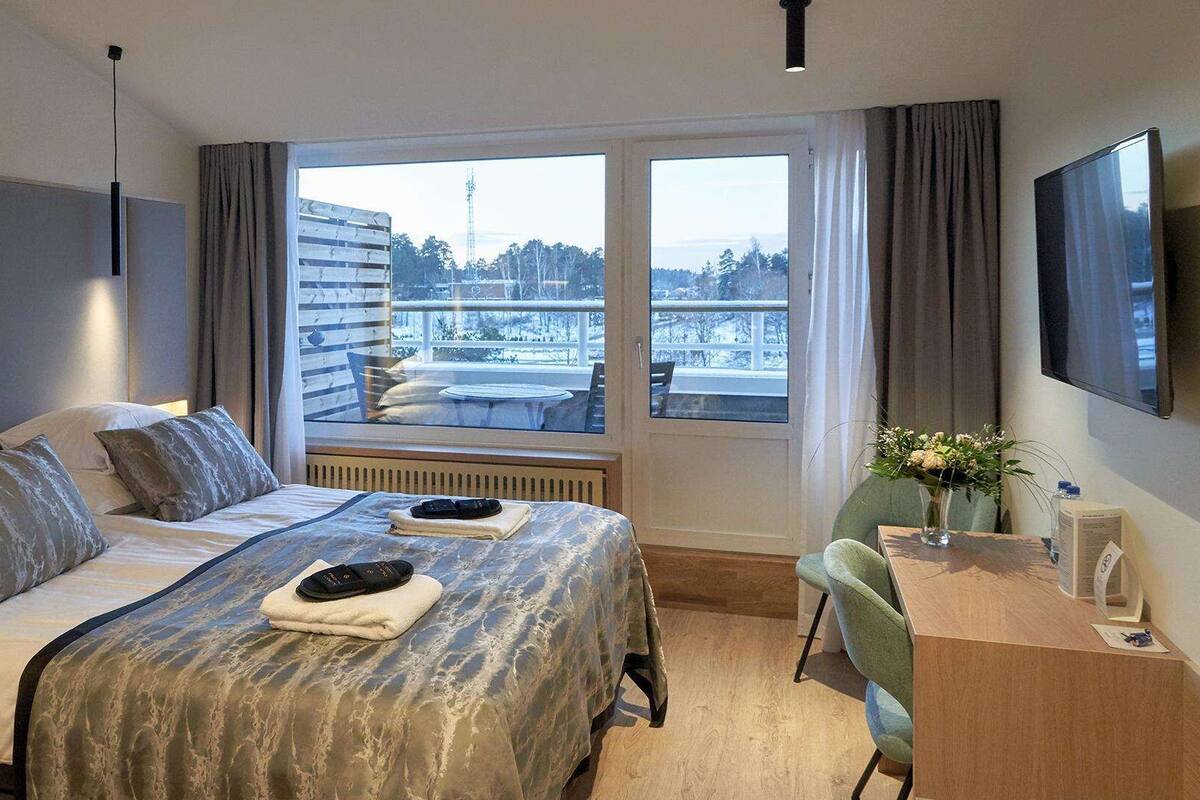 A hotel room with a large bed, a desk, and a view of a cityscape through the window.