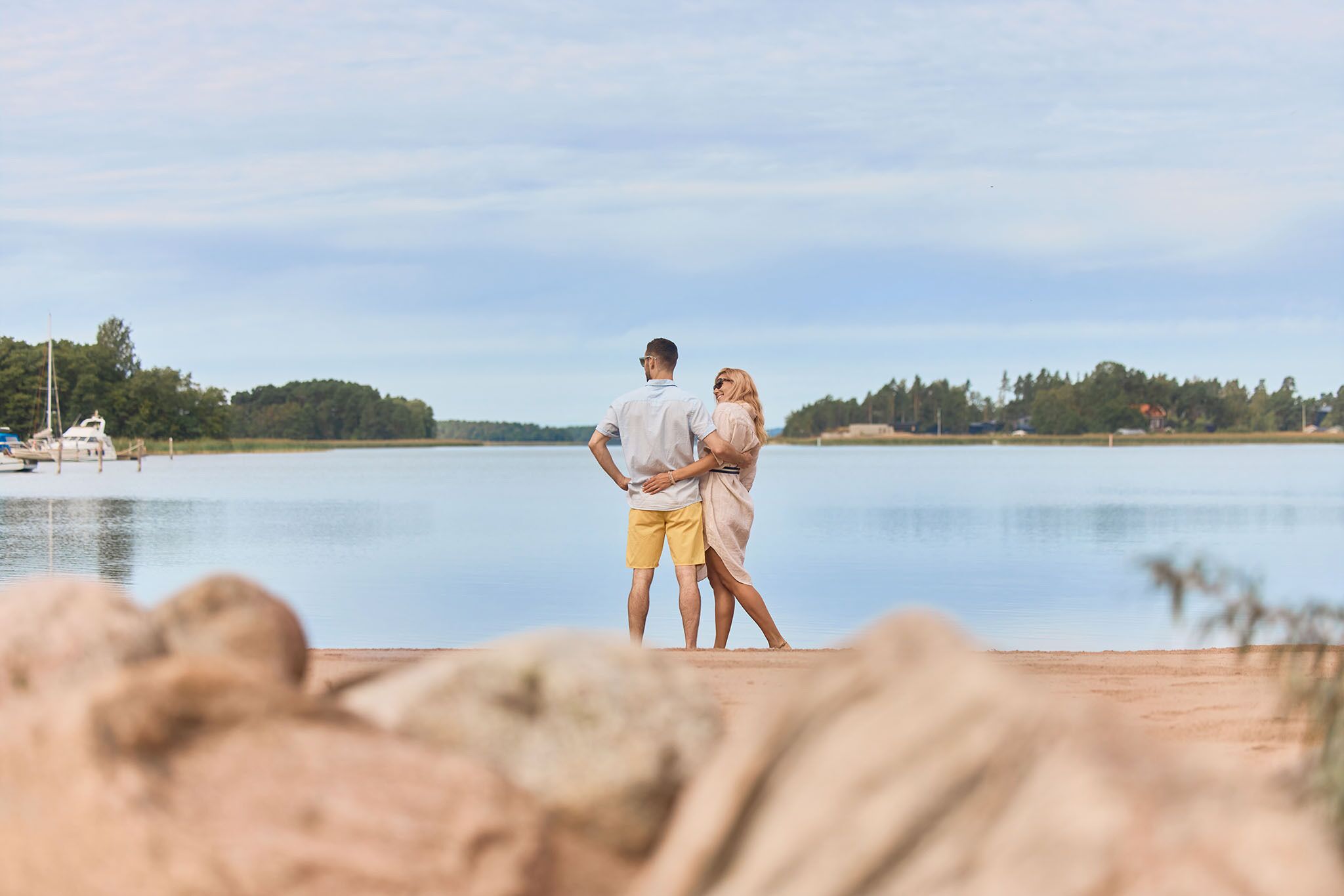 A couple standing on a sandy beach with a lake and boats in the background.