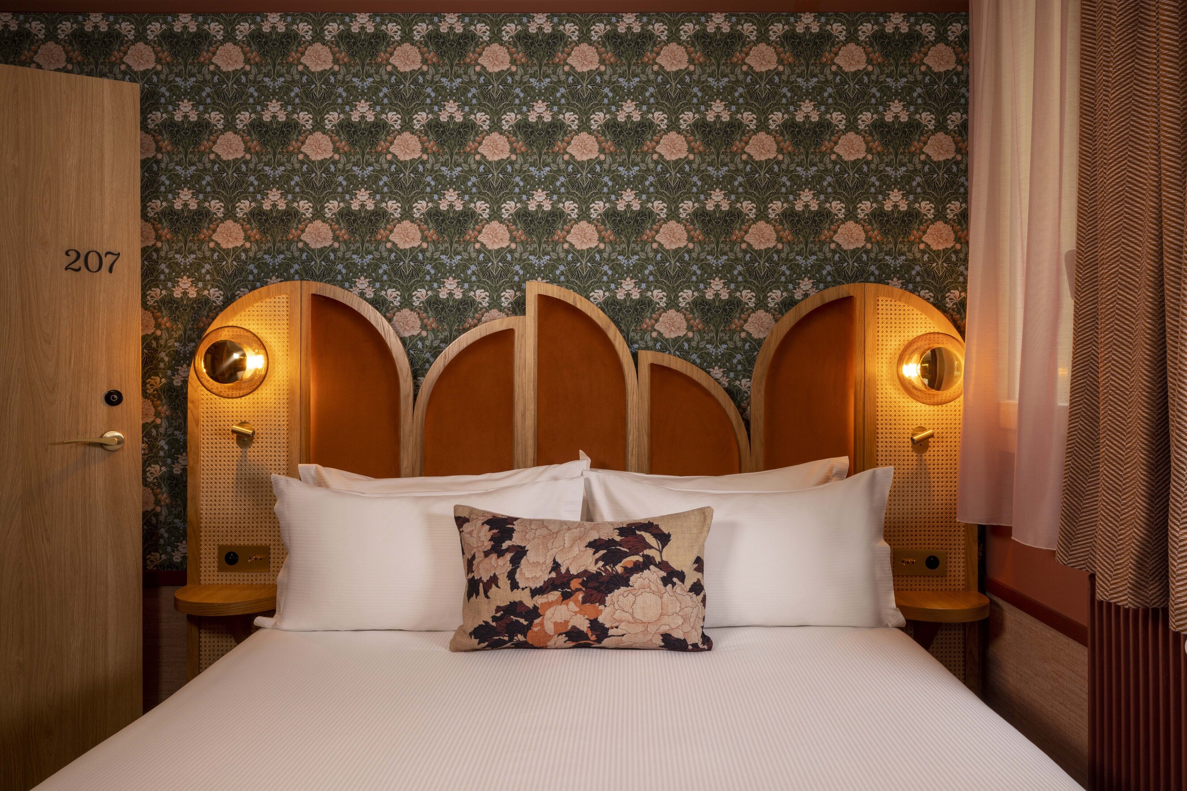 Habitación de hotel con cama, papel tapiz floral, una puerta de madera y el número 207 en la puerta.