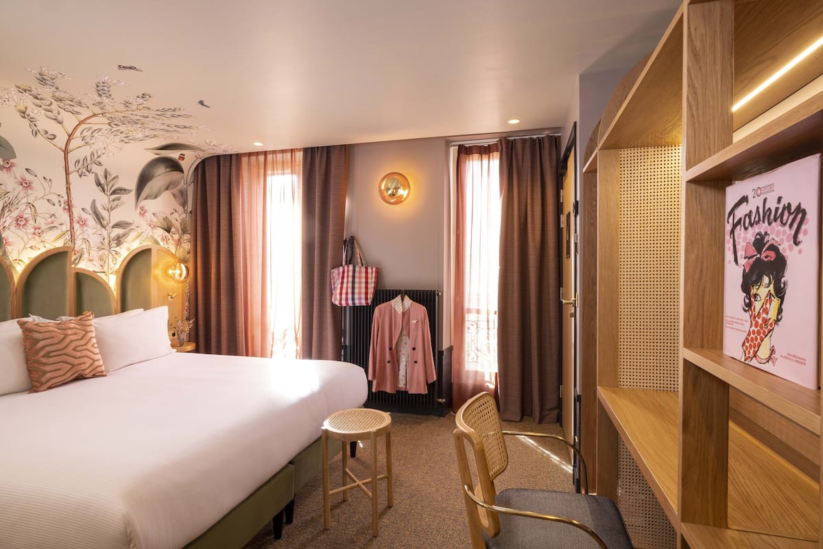 Een hotelkamer met een groot bed, een klein rond tafeltje, een stoel, een kapstok met een jas, en een muur met een poster.