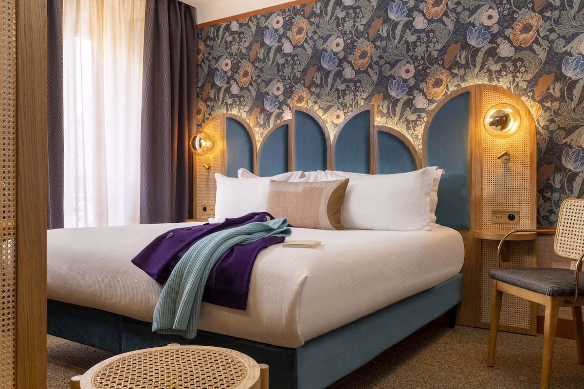 Een net opgemaakt bed met een blauw hoofdbord, een houten nachtkastje en een rieten stoel.