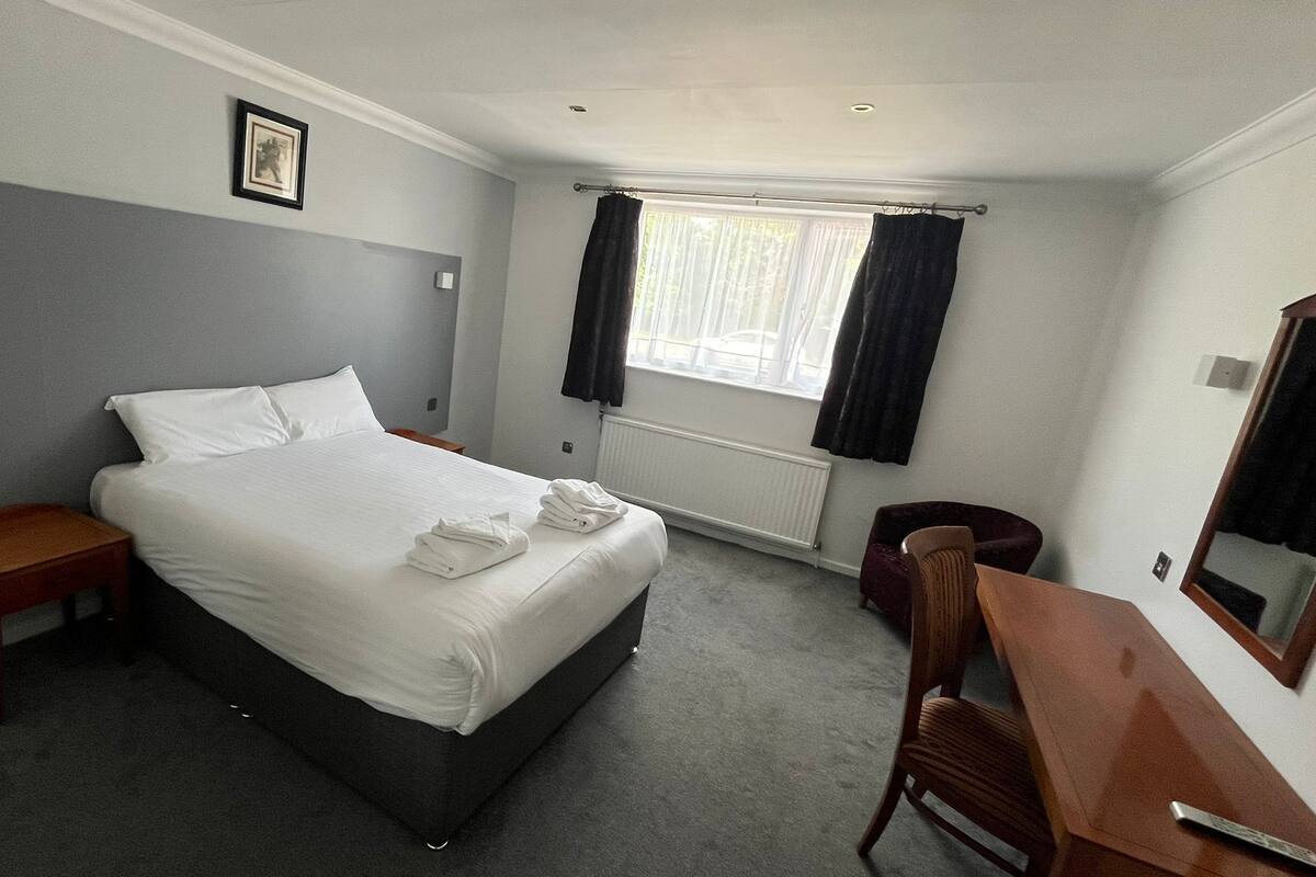 A hotel room with a bed, a desk, a chair, a window with curtains, a radiator, and a picture on the wall.