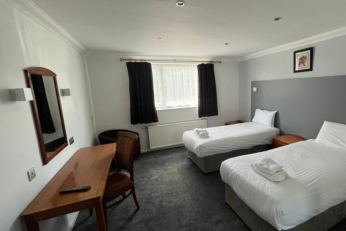 A hotel room with two single beds, a desk with a chair, a window with curtains, and a framed picture on the wall.