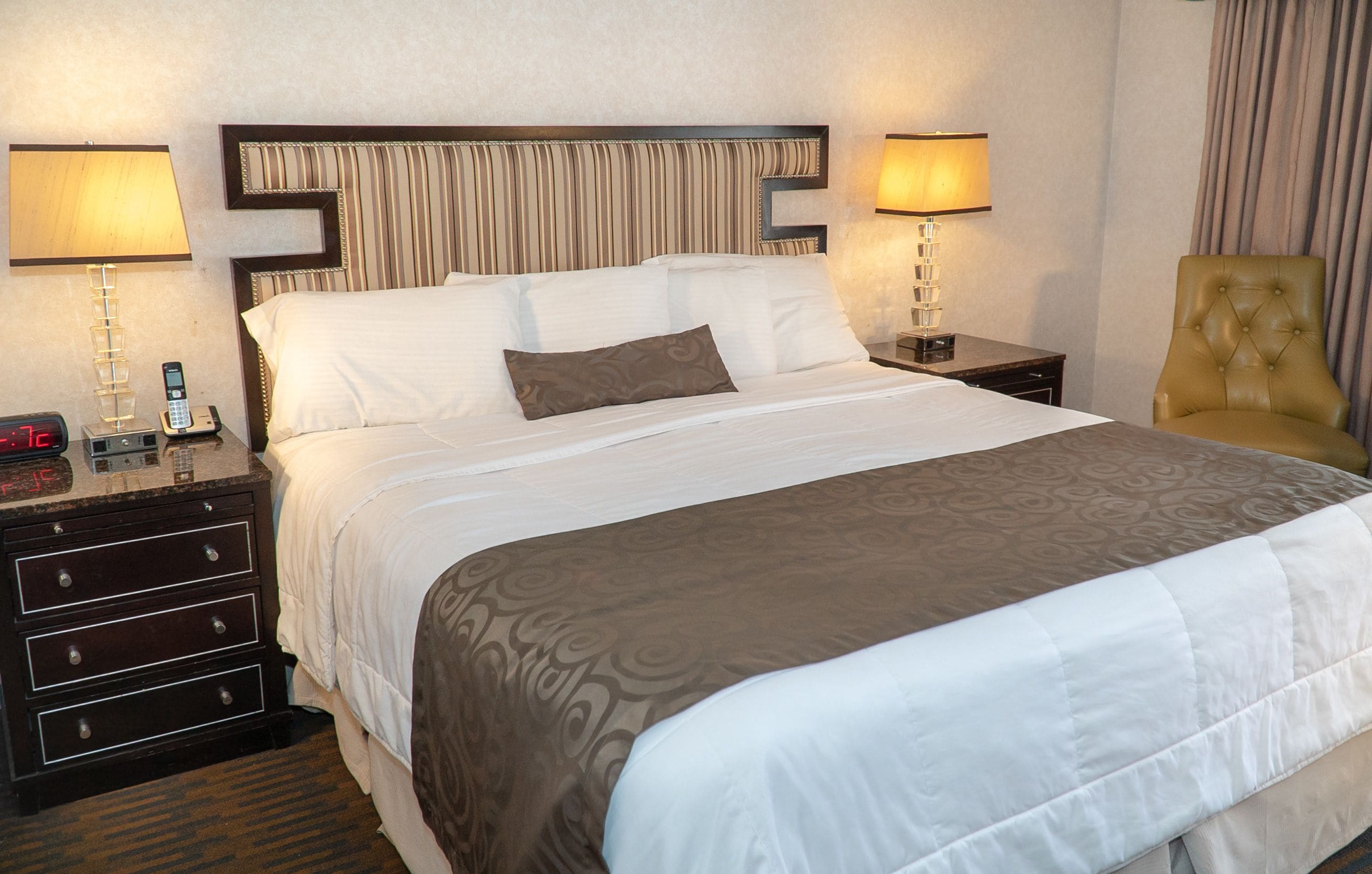 Quarto de hotel com uma cama grande, duas mesas de cabeceira com abajures, uma cadeira e um relógio.