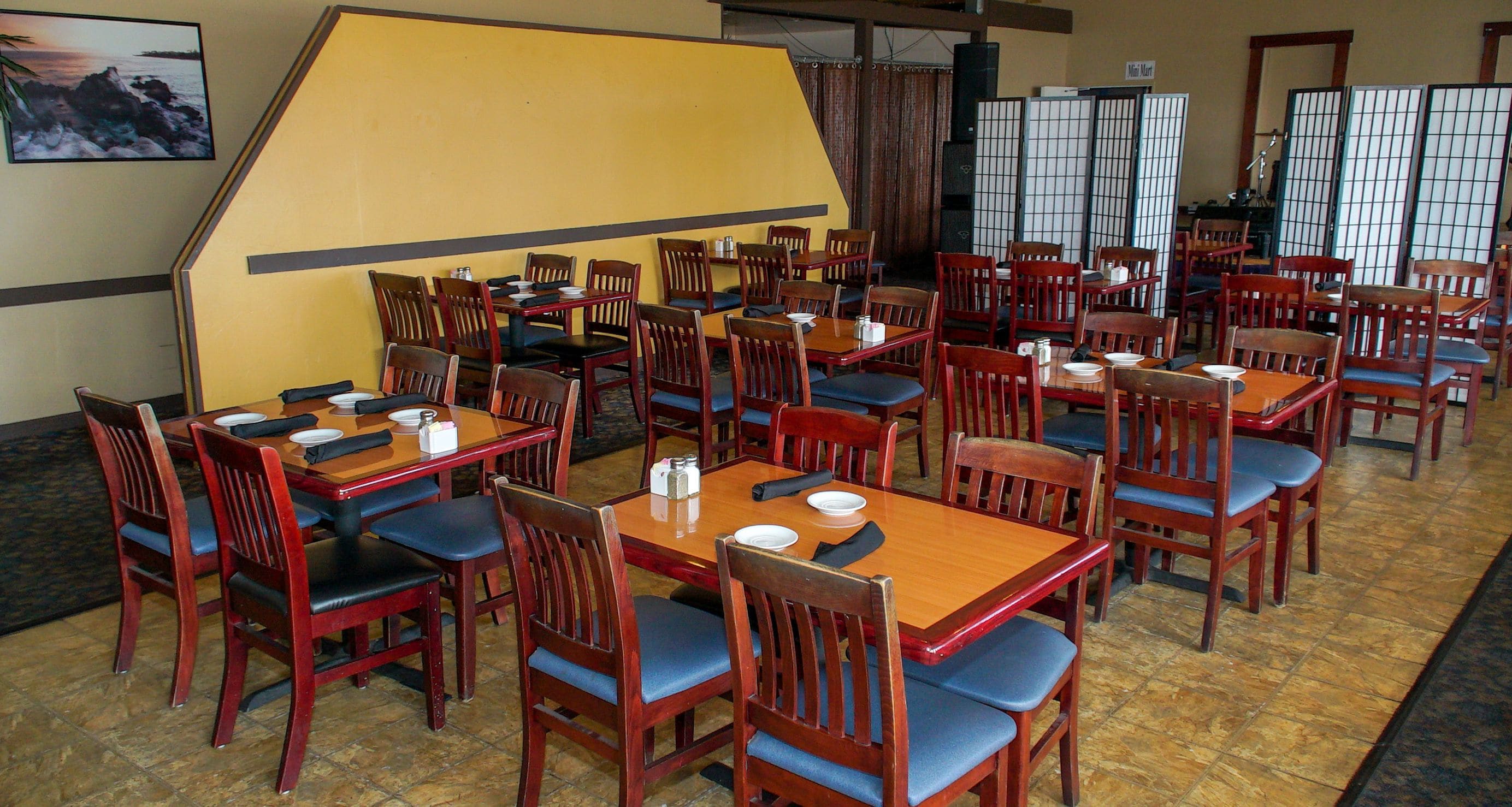 A restaurant with multiple wooden tables and chairs, a yellow wall, and a view of the sea.