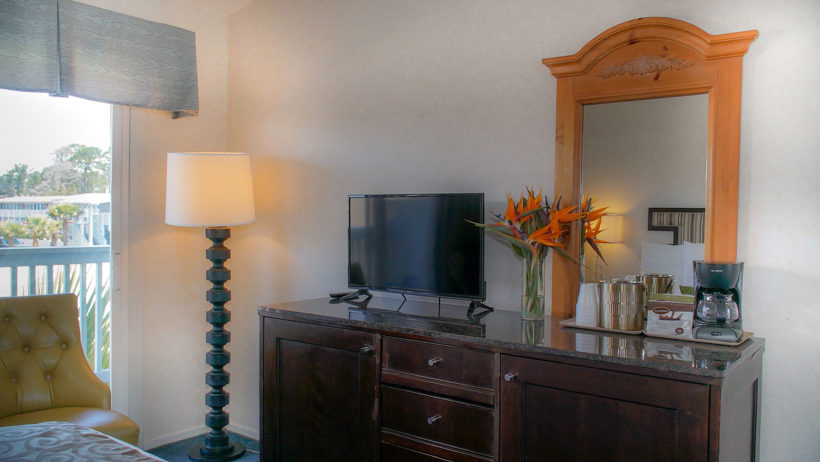 Una habitación de hotel con una cama, un televisor sobre un mueble de madera, una lámpara, un jarrón con flores y una cafetera.