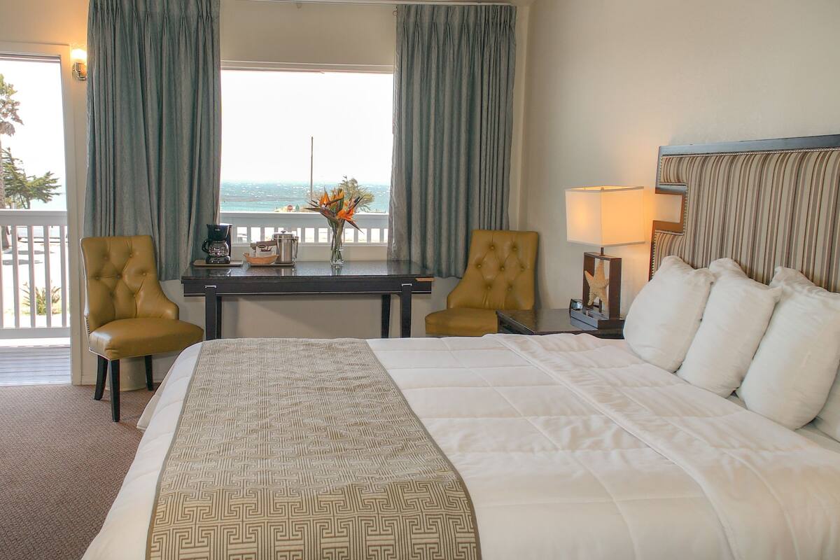 A hotel room with a large bed, two armchairs, a small table with a coffee maker, and a view of the ocean through large windows.