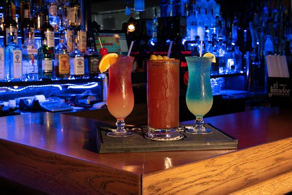 Un bar con tres bebidas en una bandeja, diversas botellas al fondo y una barra.