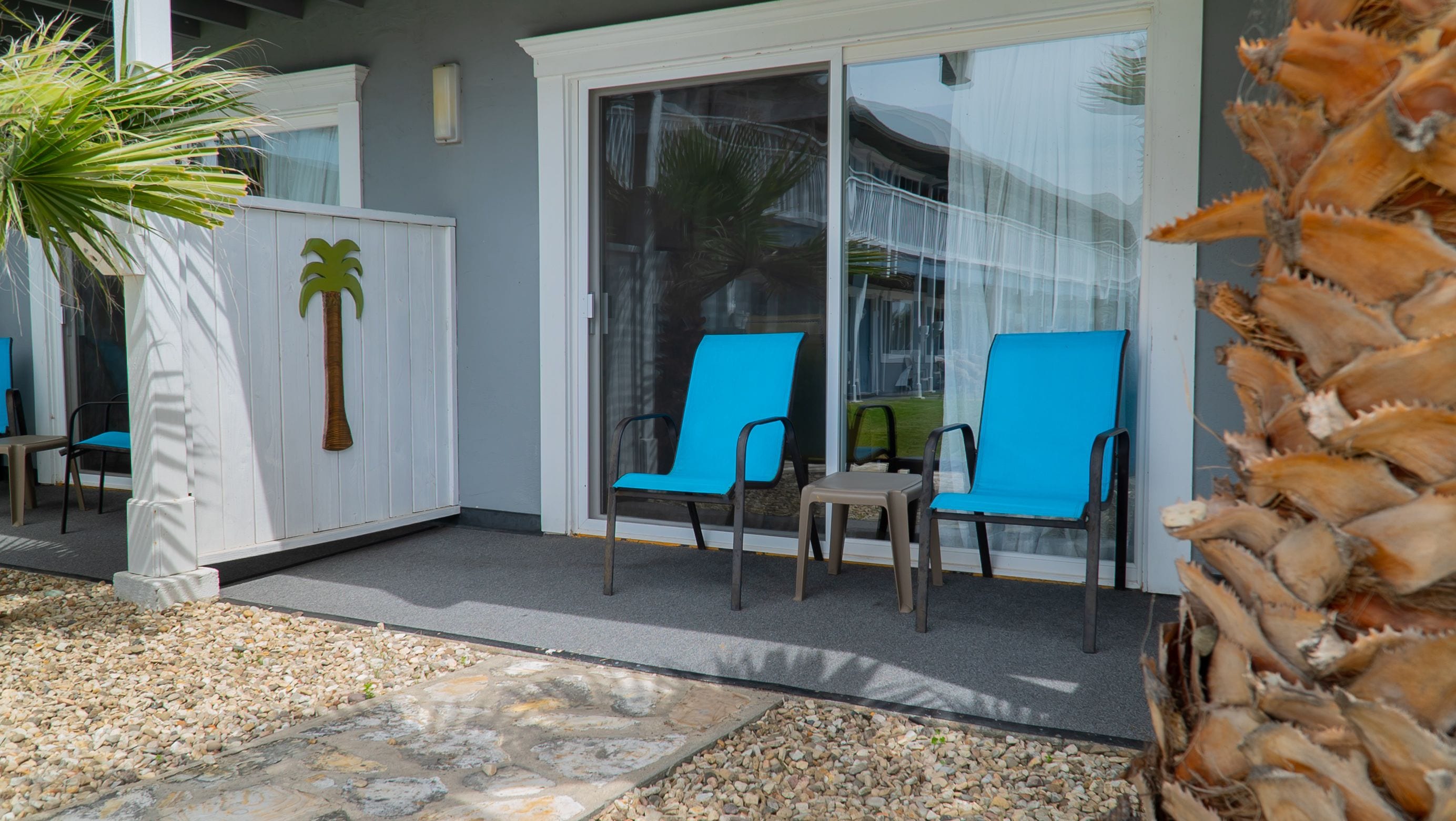 A covered outdoor seating area with two blue chairs and a table, a palm tree, and a white fence.