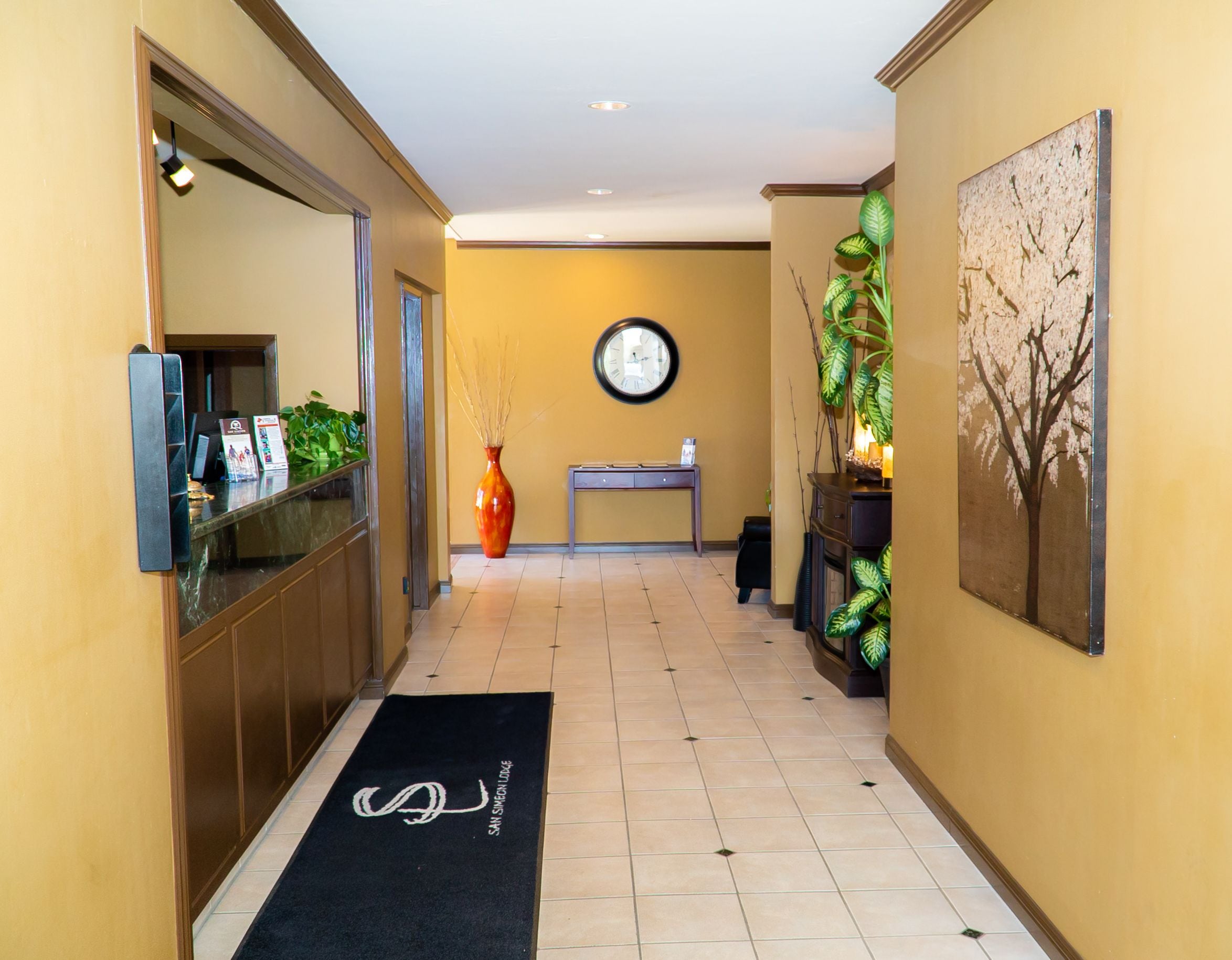 A hotel lobby with a reception desk, a clock on the wall, a black doormat with a logo, and a large painting of a tree.