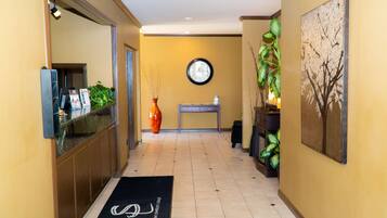 A hotel lobby with a reception desk, a clock on the wall, a black doormat with a logo, and a large painting of a tree.