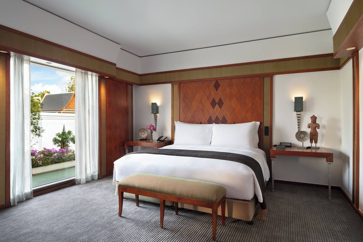 A hotel room with a large bed, a bench, a nightstand with a lamp, and a view of the outside.