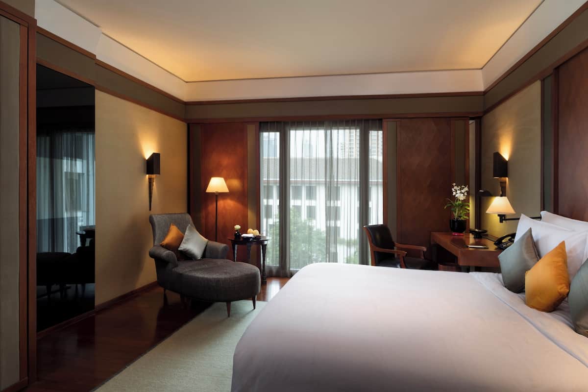 A hotel room with a large bed, a desk, a chair, and a sofa. There are two lamps on the desk and a plant on the side table.