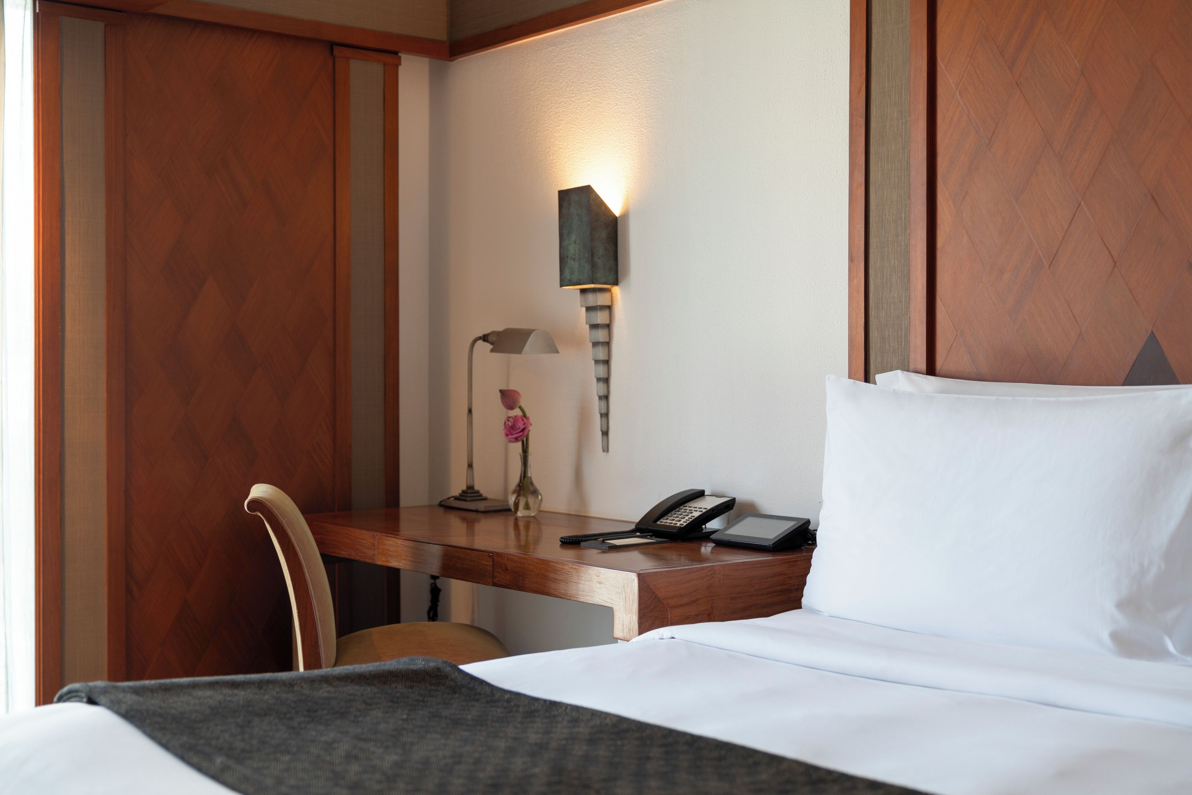 A hotel room with a wooden desk, a chair, a bedside table with a lamp, a phone, and a bed with white bedding.