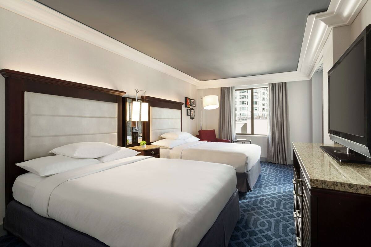 A hotel room with two beds, a TV, a desk, and a window with a view of buildings.