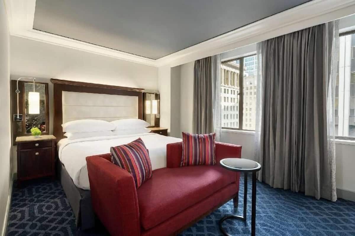 A hotel room with a bed, a red sofa, a small round table, a nightstand, a mirror, and a window with curtains.