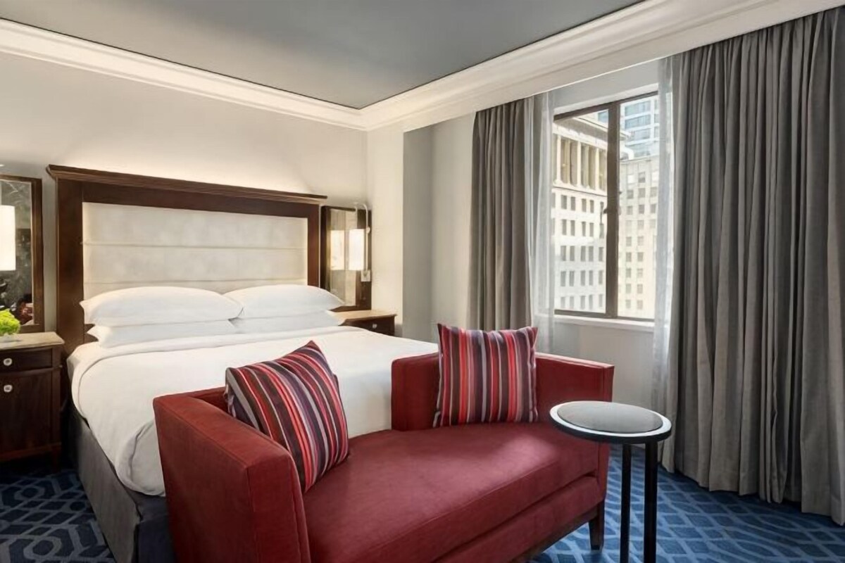 A hotel room with a bed, a red sofa, a small round table, and a window with curtains.