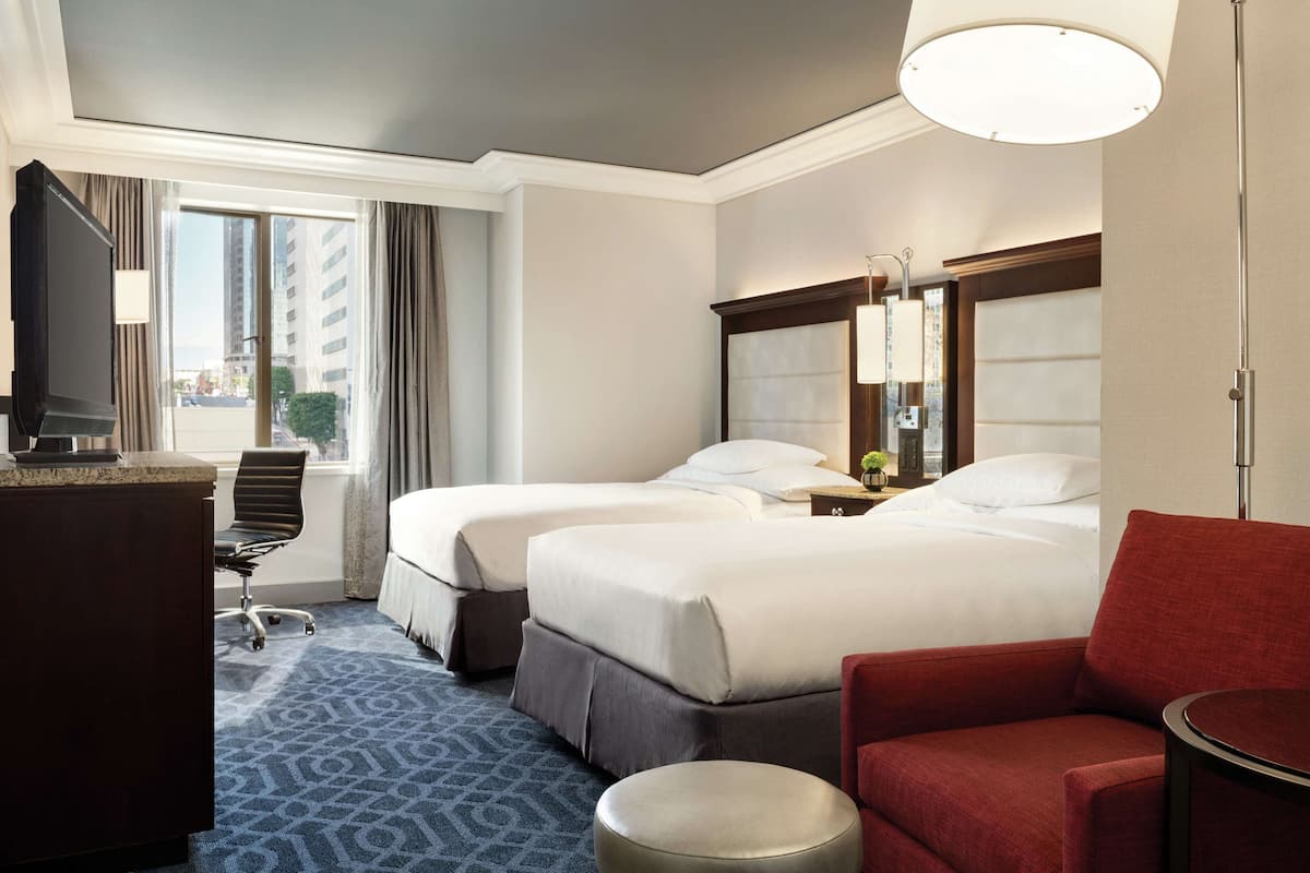 A hotel room with two beds, a desk, a chair, a red armchair, and a lamp.