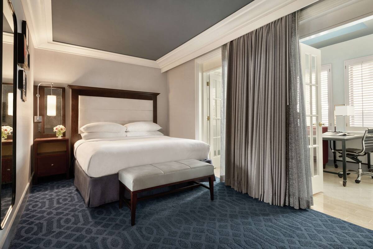 A hotel room with a large bed, a nightstand with a lamp, a desk with a chair, and a view of the outside through a window.