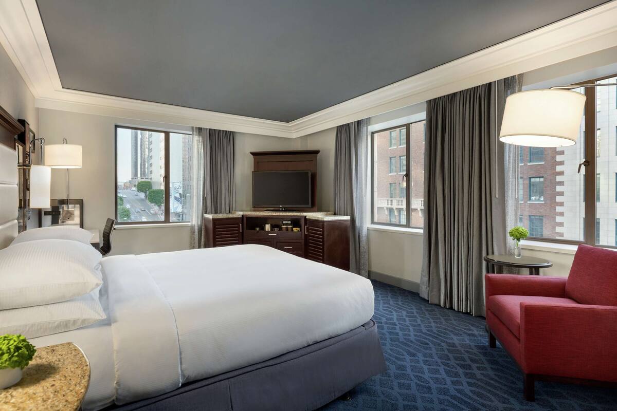 A hotel room with a large bed, a desk with a flat-screen TV, a red armchair, and a view of the city through large windows.
