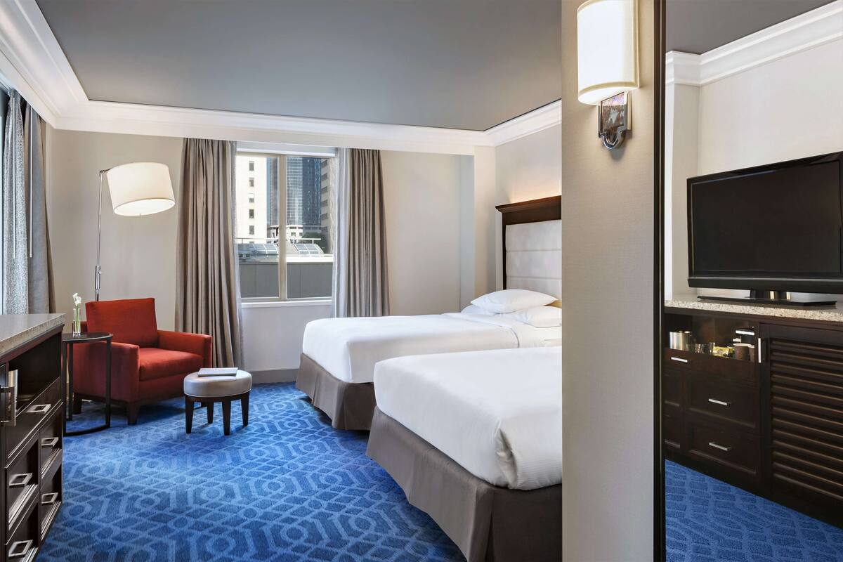 A hotel room with two beds, a TV, a desk with a chair, and a window with curtains.