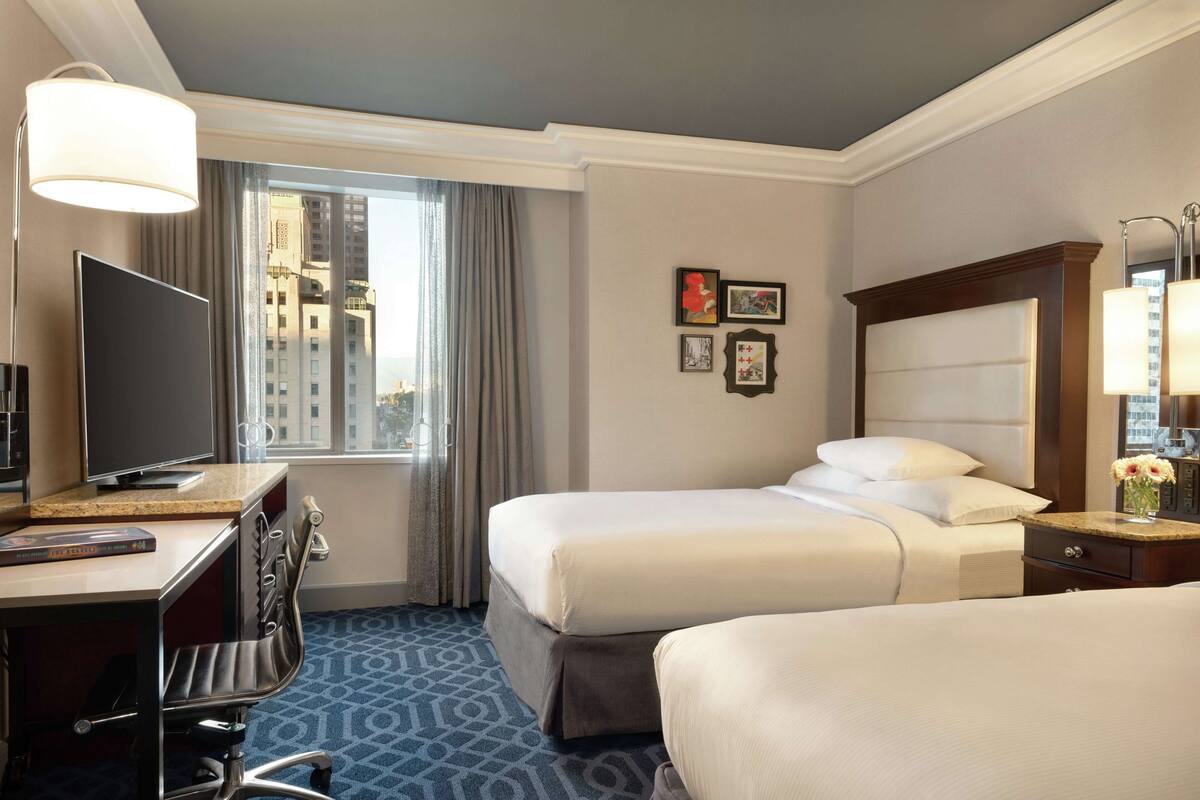 A hotel room with two beds, a desk with a TV, a chair, a window with curtains, and framed pictures on the wall.