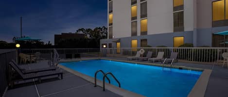 A swimming pool with lounge chairs and a building with illuminated windows in the background.