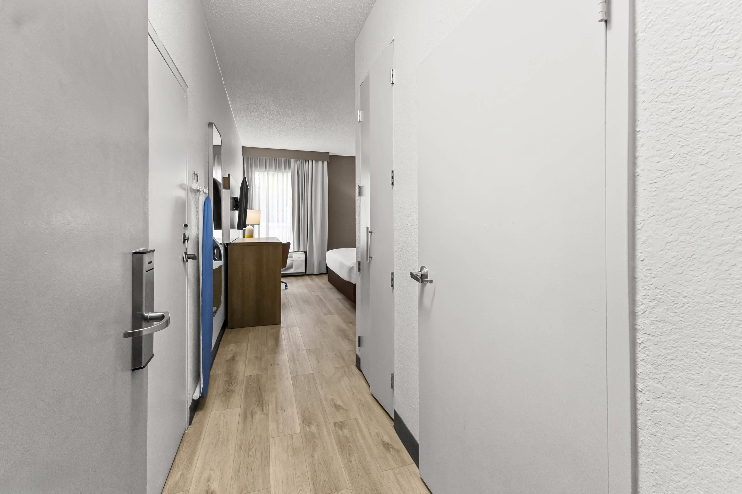 A narrow hallway with a wooden floor leading to a room with a bed and a window with curtains.