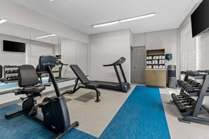 Uma academia com equipamentos de ginástica, um espelho, uma televisão e uma porta para o vestiário.