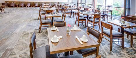 A spacious restaurant with multiple dining tables and chairs, large windows offering a view of the outdoors, and a carpeted floor.