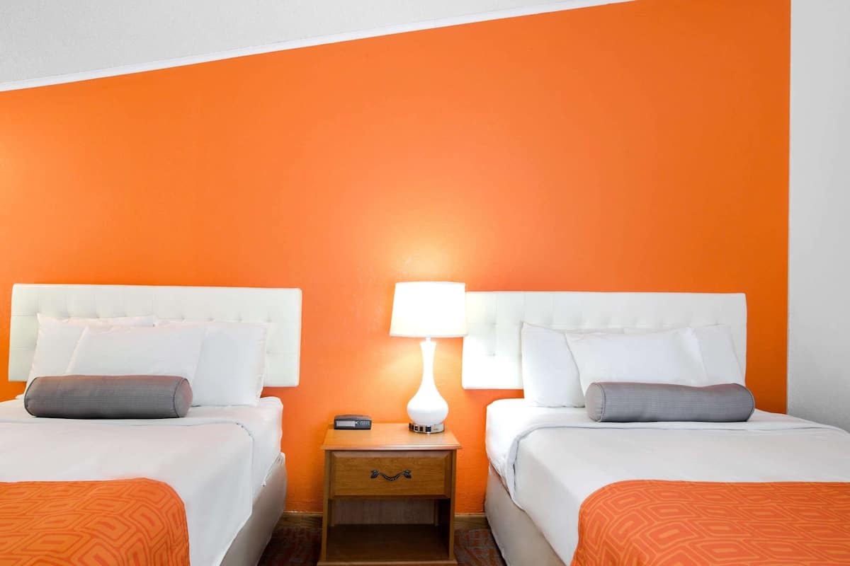 A hotel room with two beds, each with white bedding and orange throw pillows. There is a nightstand with a lamp and a remote control between the beds.
