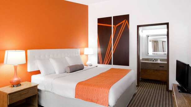 Habitación de hotel con cama, mesita de noche, televisor, espejo y baño con lavamanos y espejo.