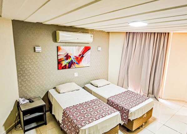 A hotel room with two single beds, an air conditioning unit, a painting on the wall, a bedside table with a phone and charger, and a window with curtains.