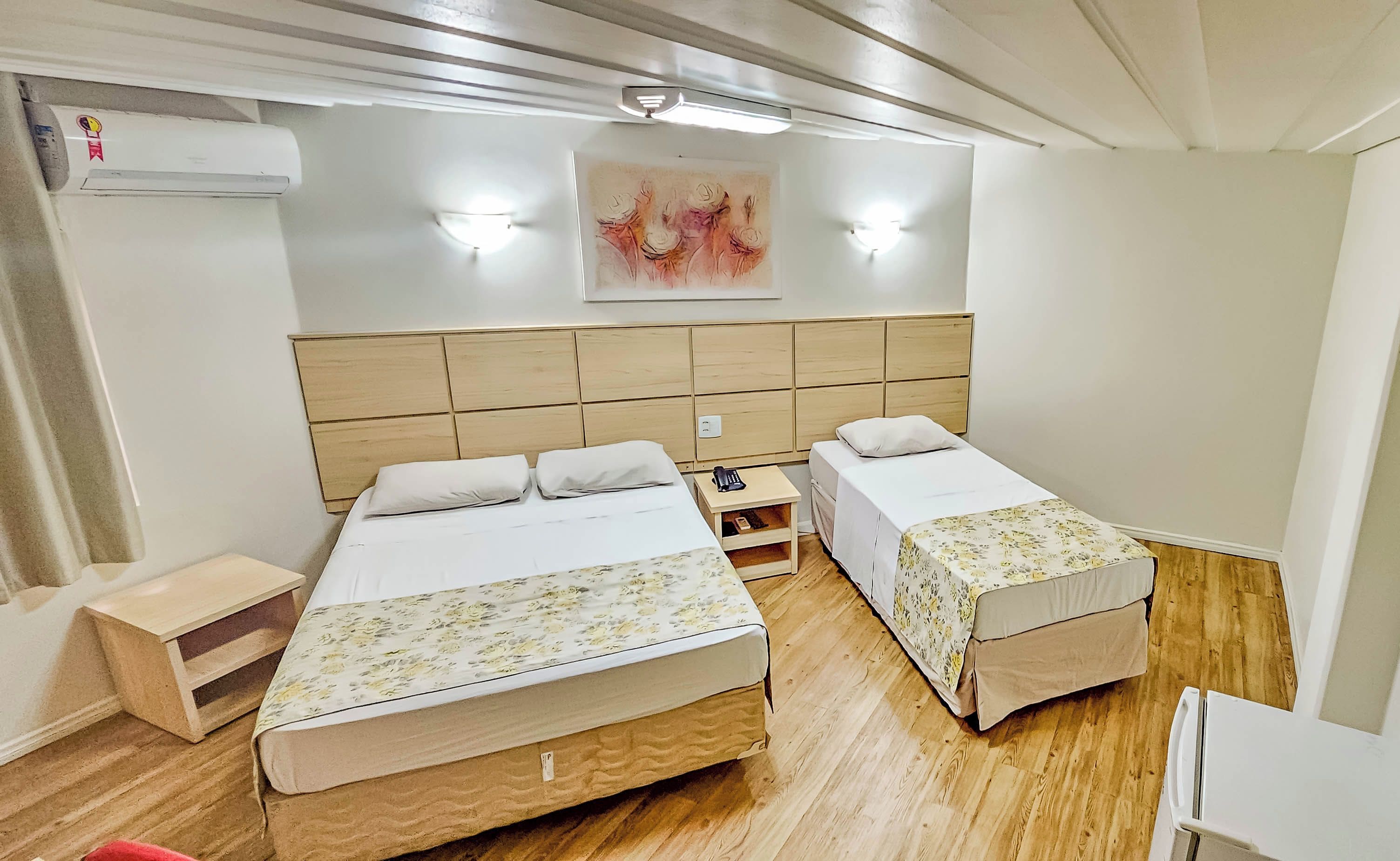 A hotel room with two beds, a wooden headboard, a small bedside table, a wall-mounted air conditioner, and a painting.