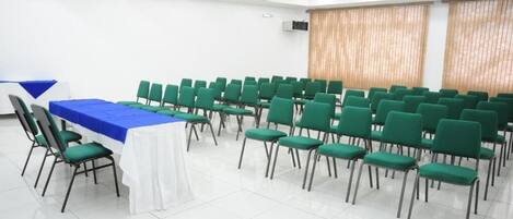 A conference room with rows of green chairs, a white table with a blue tablecloth, and a large window with vertical blinds.