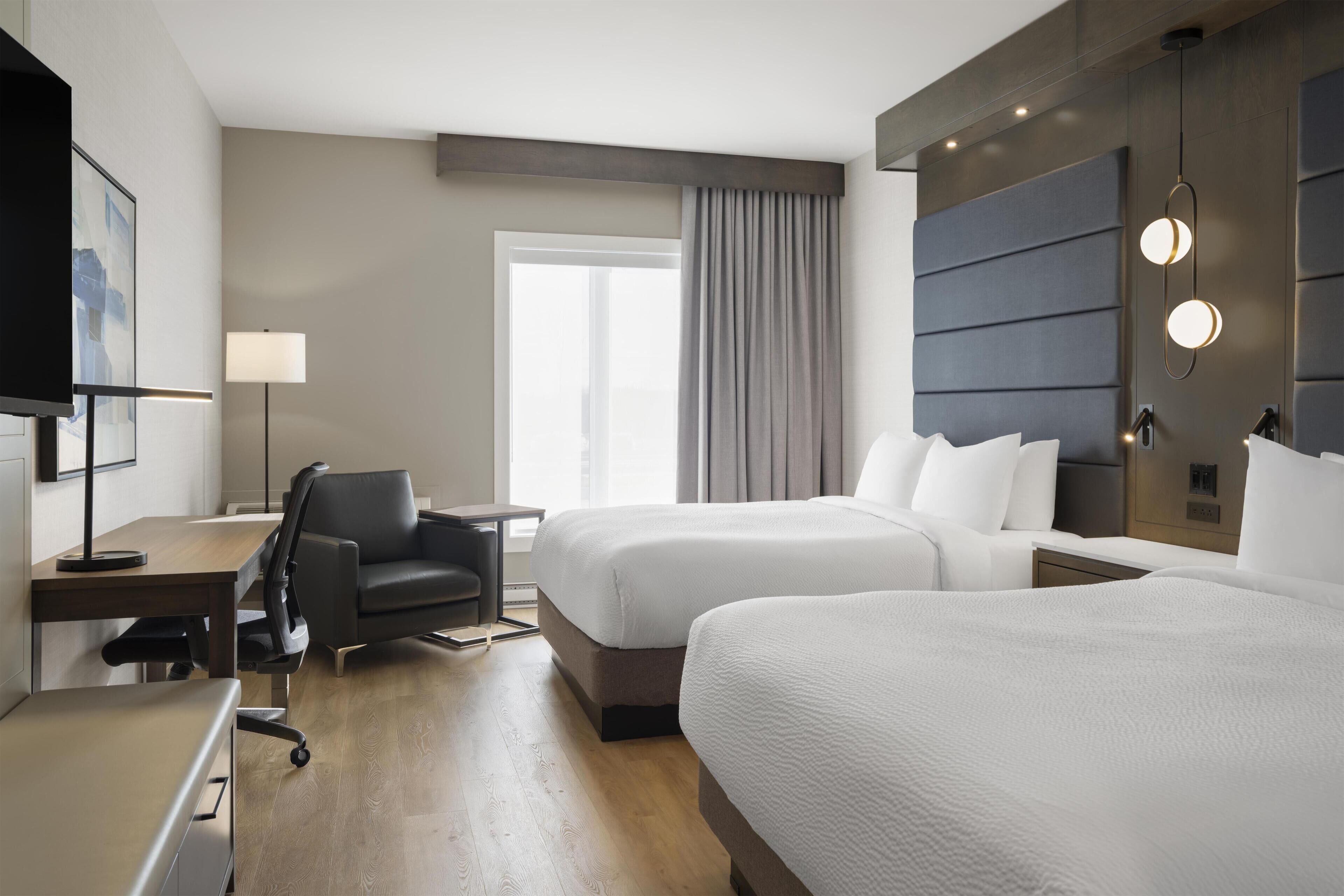 Habitación de hotel con dos camas, un escritorio, una silla, un televisor, una lámpara y un ventanal grande con cortinas.