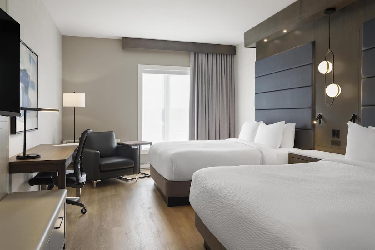 A hotel room with two beds, a desk, a chair, a TV, a lamp, and a large window with curtains.