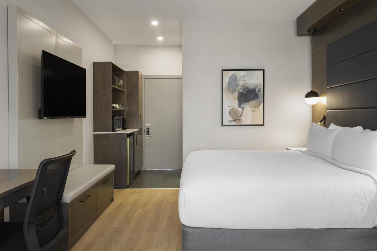 A modern hotel room with a bed, a desk, a television, and a painting on the wall.