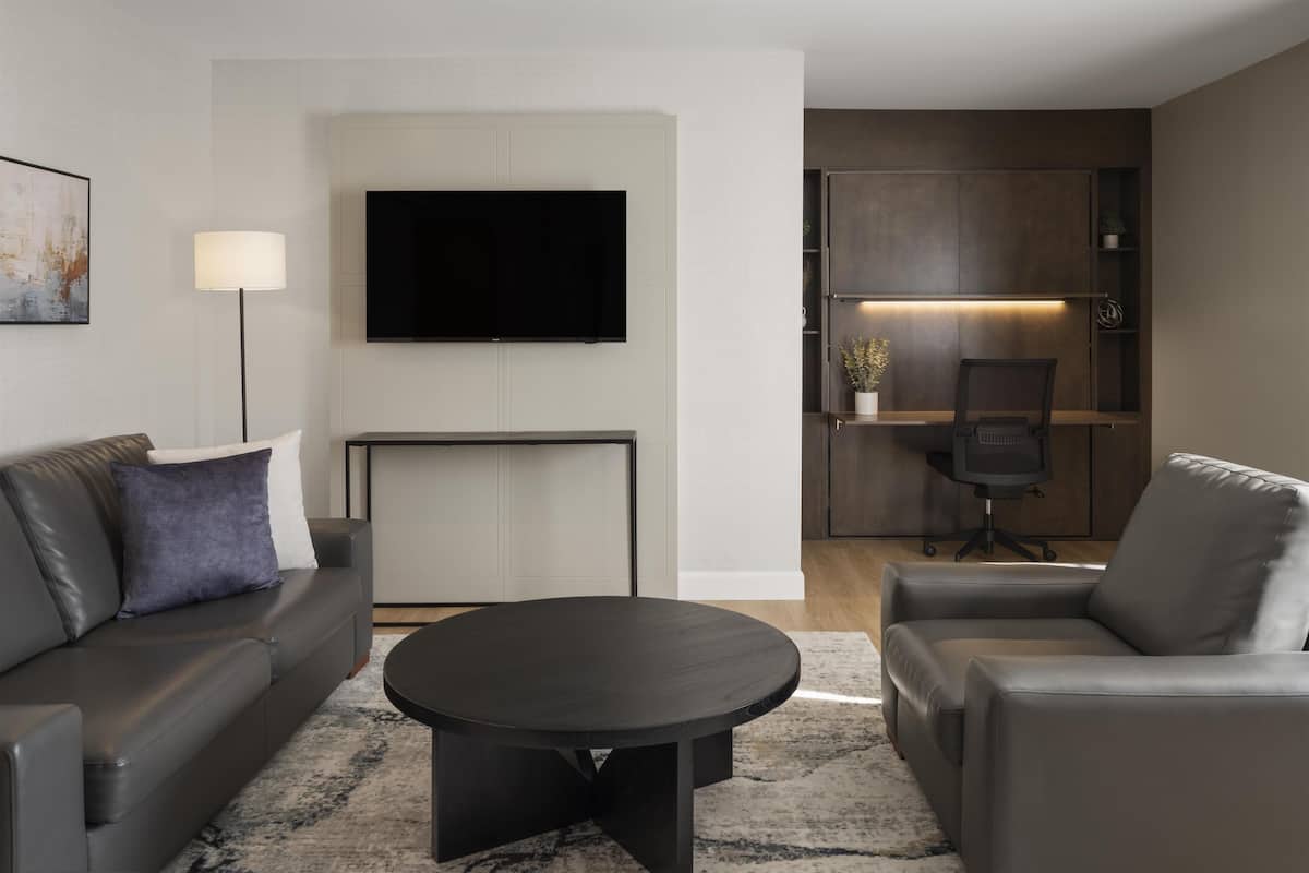 A modern living room with a sofa, armchair, and a round coffee table. A flat-screen TV is mounted on the wall, and there is a desk with a chair in the background.