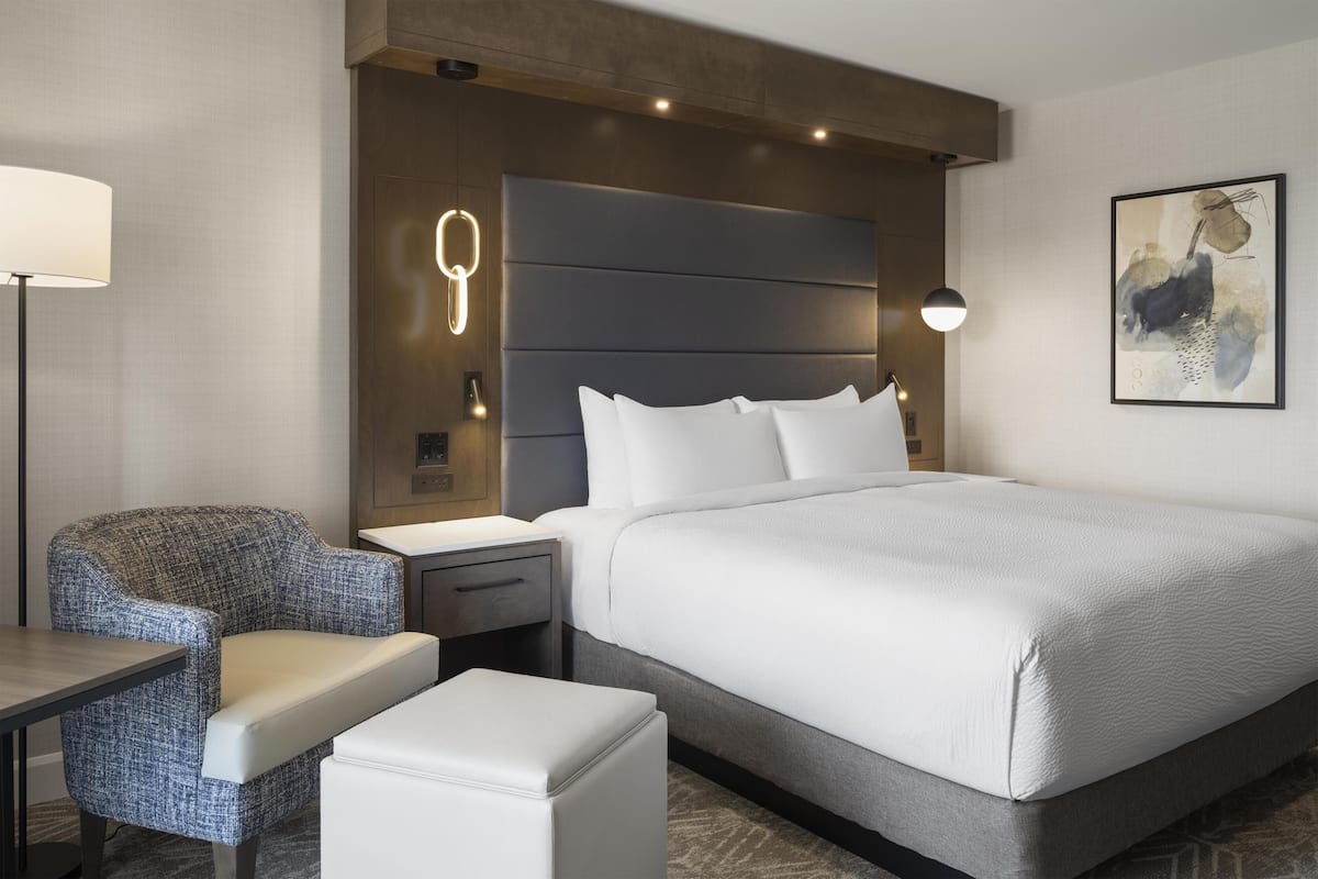 A modern hotel room with a large bed, a seating area with a chair and ottoman, a bedside table, and a wall-mounted lamp.