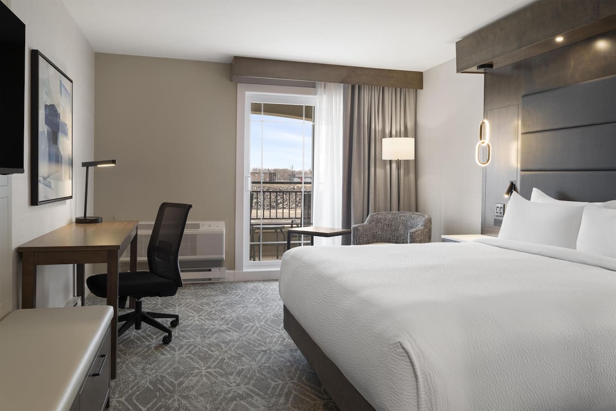 A hotel room with a large bed, a desk, a chair, a lamp, and a view of the city through a balcony.