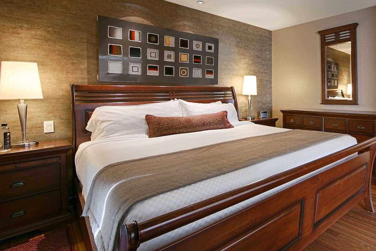 A hotel room with a large bed, two bedside tables, a nightstand, a dresser, a mirror, and a wall art piece.