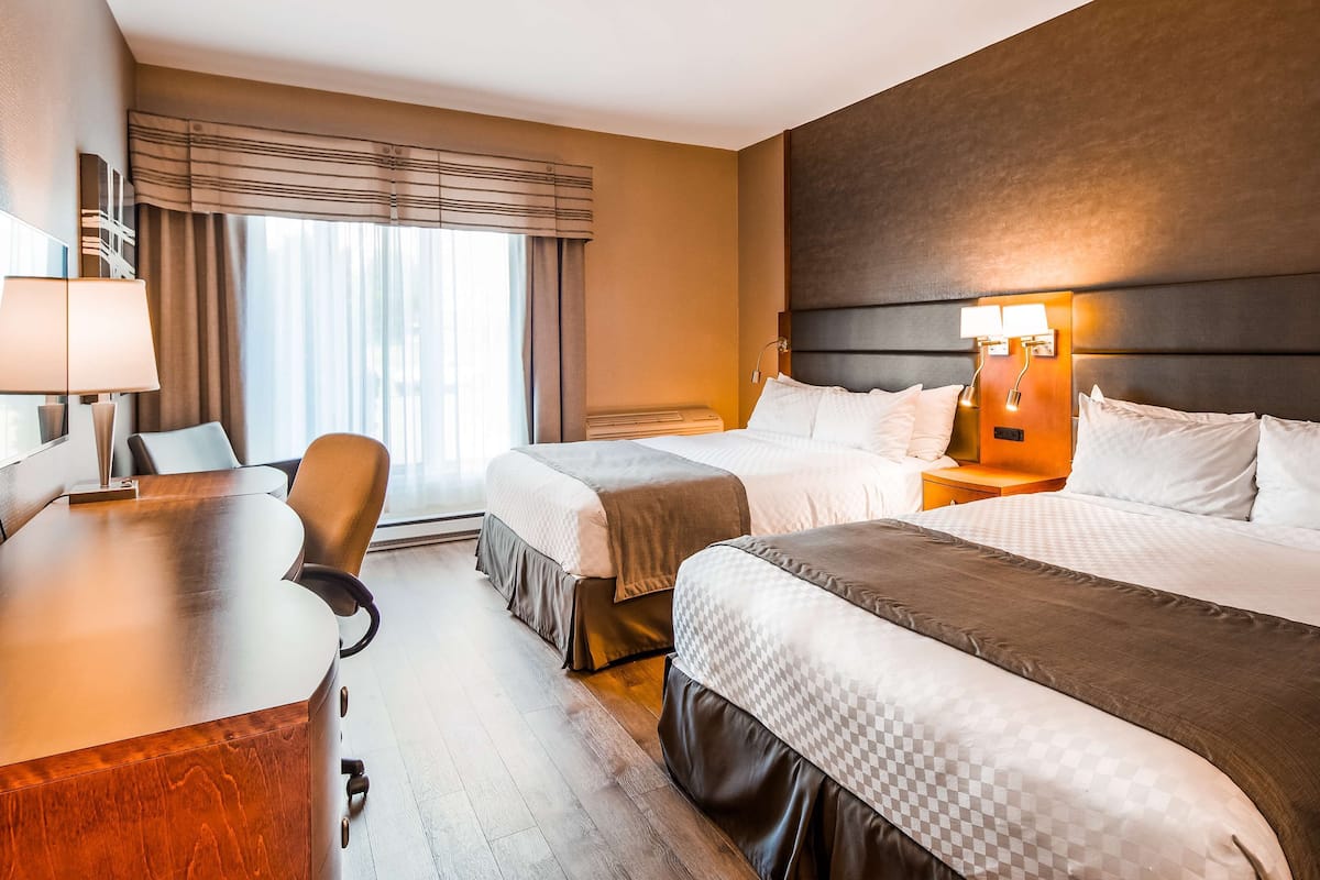 A hotel room with two beds, a desk, a chair, a lamp, and a window with curtains.