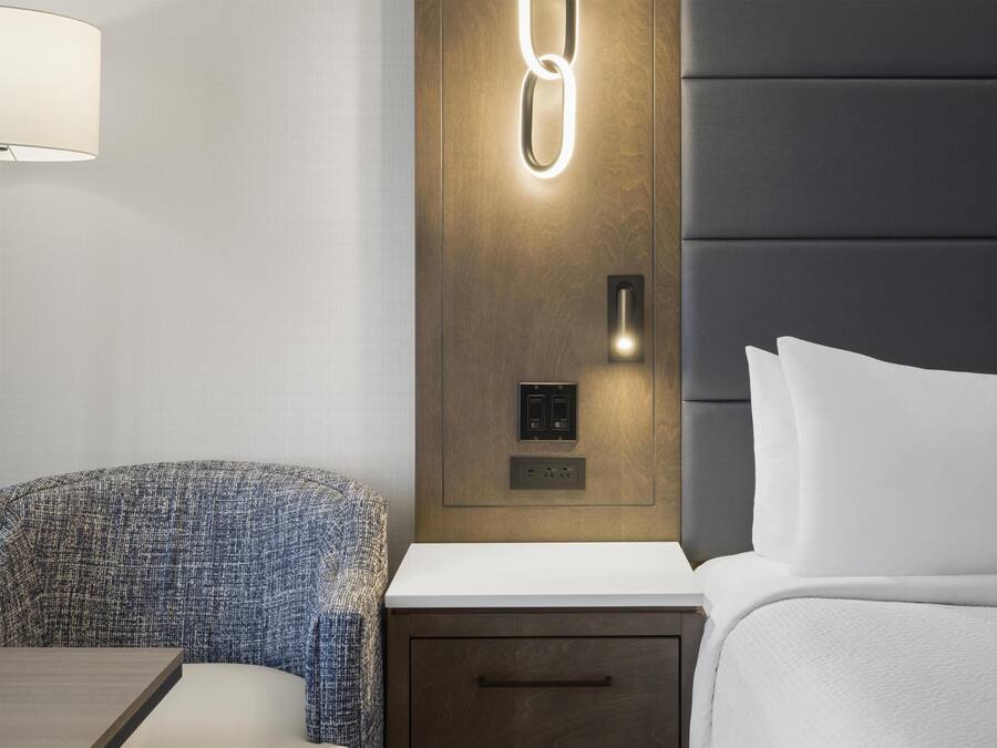 A hotel room with a bed, bedside table, lamp, and a wall-mounted light fixture.