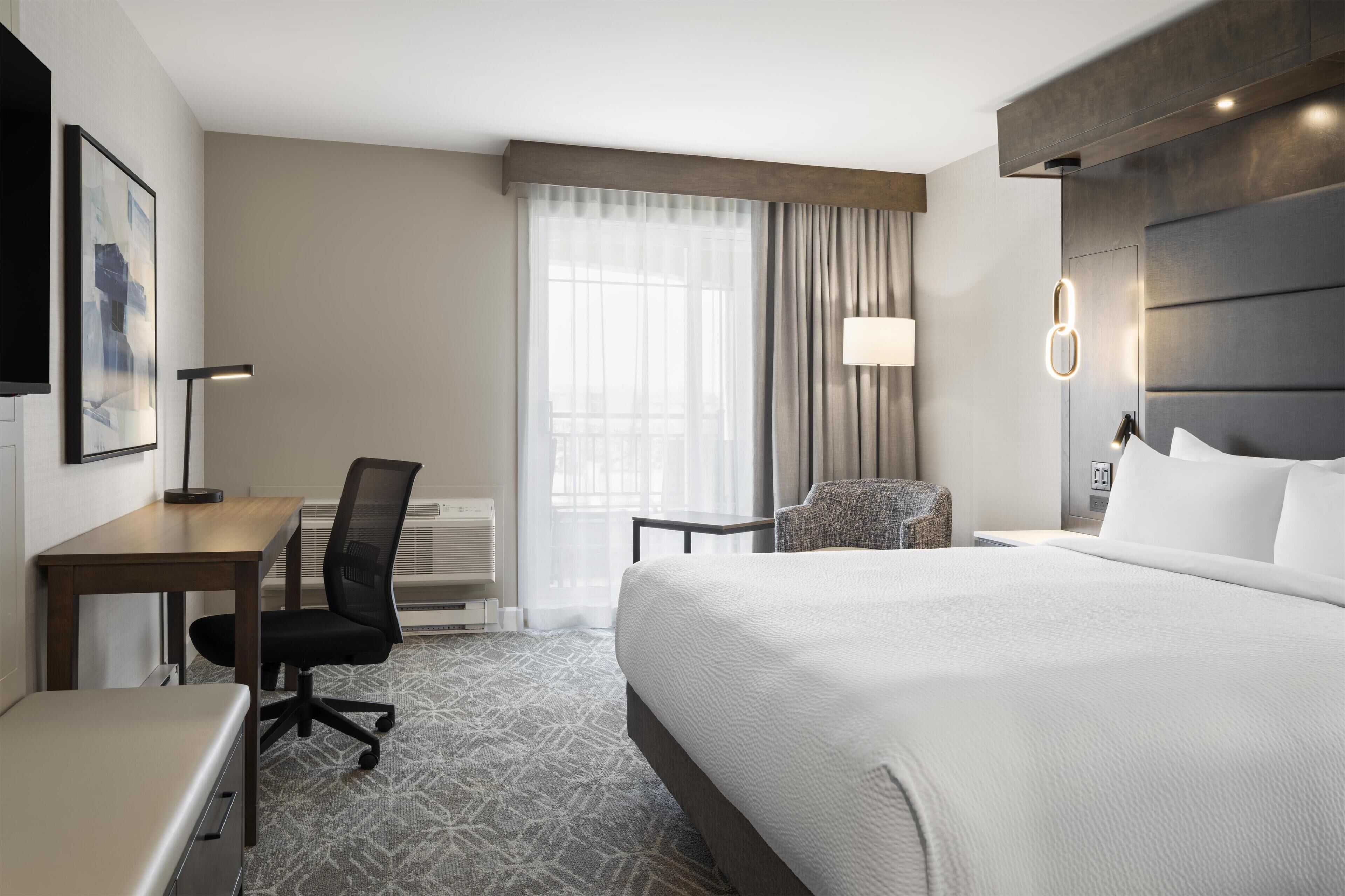 Une chambre d’hôtel avec un lit, un bureau, une chaise et une grande fenêtre avec des rideaux.