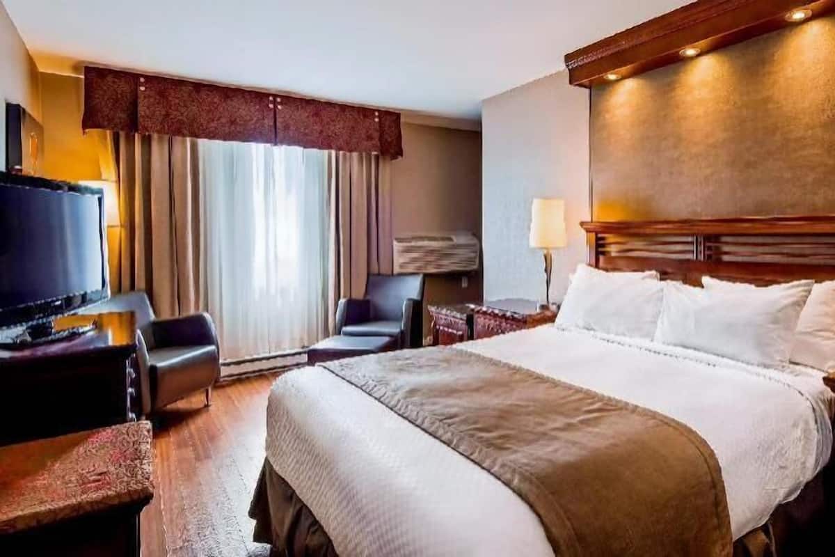 A hotel room with a large bed, a desk with a flat-screen TV, a chair, and a desk lamp.